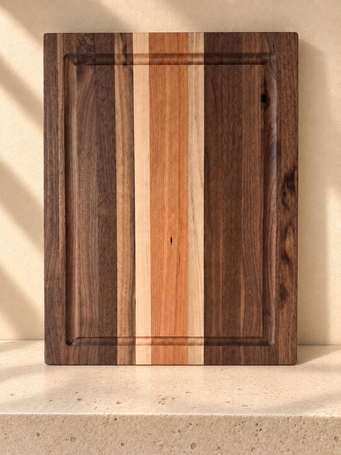 The Hawthorne hardwood cutting board with contrasting wood stripes and a juice groove, displayed on warm cream stone.